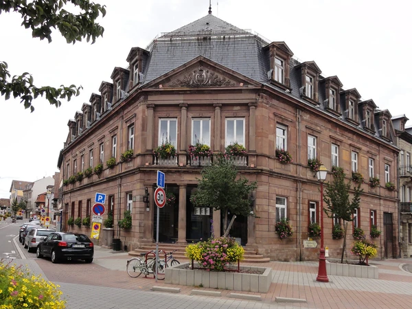 Photographie professionnelle d'une rue typique de Schiltigheim (Alsace) avec bâtiments résidentiels ou commerciaux multi-étages d'architecture locale, photographiée de jour. La scène doit montrer clairement un environnement urbain/périurbain du Grand Est, avec une bonne profondeur de champ et une composition équilibrée. L'image doit transmettre une impression de proximité locale et d'ancrage territorial, complémentaire à l'image d'immeuble déjà utilisée. Pas de personnes, véhicules ou éléments anecdotiques en avant-plan.